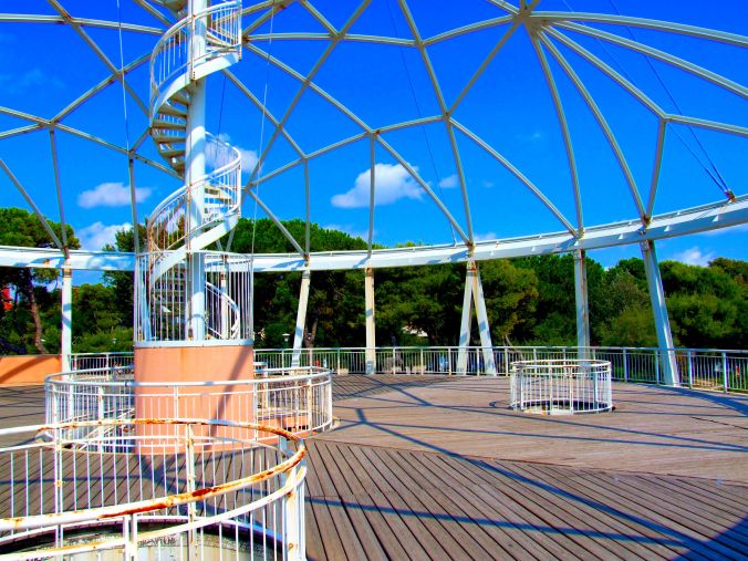 Blue Moon, Piazzale Bucintoro 1 – 30126 Lido Di Venezia. Shadow triangles move slowly across the wooden decking as the sun moves across the sky. The ‘Blue Moon’ was designed by Giancarlo de Carlo (1919-2005), professor emeritus at the Venice School of Architecture (Università Iuav di Venezia), founded in 1926 as the Istituto Universitario di Architettura di Venezia. The building project began in 1995 and the main construction was completed by 2002. It stands at the intersection between the Gran Viale Santa Maria Elisabetta (a road that crosses the Lido from the point where passengers from Venice disembark) and the coast road that goes from the Murazzi to the sand dunes of San Nicolò and serves the spiaggia comunale (public beach) at the northern end of the Lido. https://witness.theguardian.com/assignment/55718213e4b019d982cba15c/1563276
